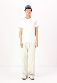 White crew-neck t-shirt, beige wide-leg trousers, gray and white sneakers, blue ribbed beanie, standing on a plain background.