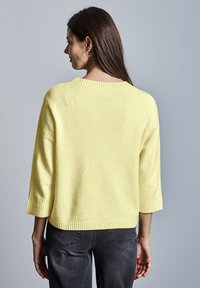 Gelber Strickpullover mit Dreiviertelärmeln, geripptem Halsausschnitt und Saum. Getragen mit dunkelgrauen Jeans. Weicher Stoff, lässiges Design.