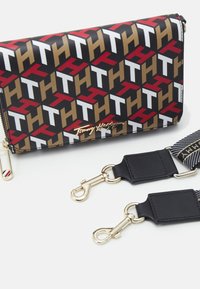 Czarny portfel z geometrycznym wzorem w kolorze czerwonym, białym, złotym i brązowym. Posiada złote logo i wyposażony jest w odczepiane czarne paski.