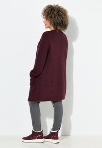 Maglione lungo color bordeaux con lavorazione a maglia testurizzata, due tasche laterali e una vestibilità comoda, abbinato a jeans grigi slim e stivaletti rossi alla caviglia.