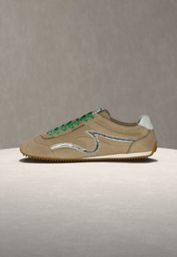 Μπεζ σουεντ αθλητικό παπούτσι με πράσινα κορδόνια, ασημί κυματιστό σχέδιο στο πλάι, λευκή ταμπέλα στη φτέρνα και καφέ λαστιχένια σόλα σε απλή επιφάνεια.