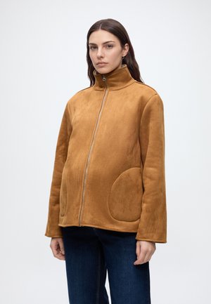 Frau trägt eine hellbraune Wildlederjacke mit Reißverschluss, hohem Kragen und abgerundeten aufgesetzten Taschen, kombiniert mit dunkelblauen Jeans, stehend vor weißem Hintergrund.