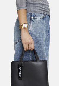 Svart läder tote bag med korta handtag och ett tag som står "LIEBESKIND." Kombination med ljusblå jeans och en grå ribbad topp. Guldur på handleden.
