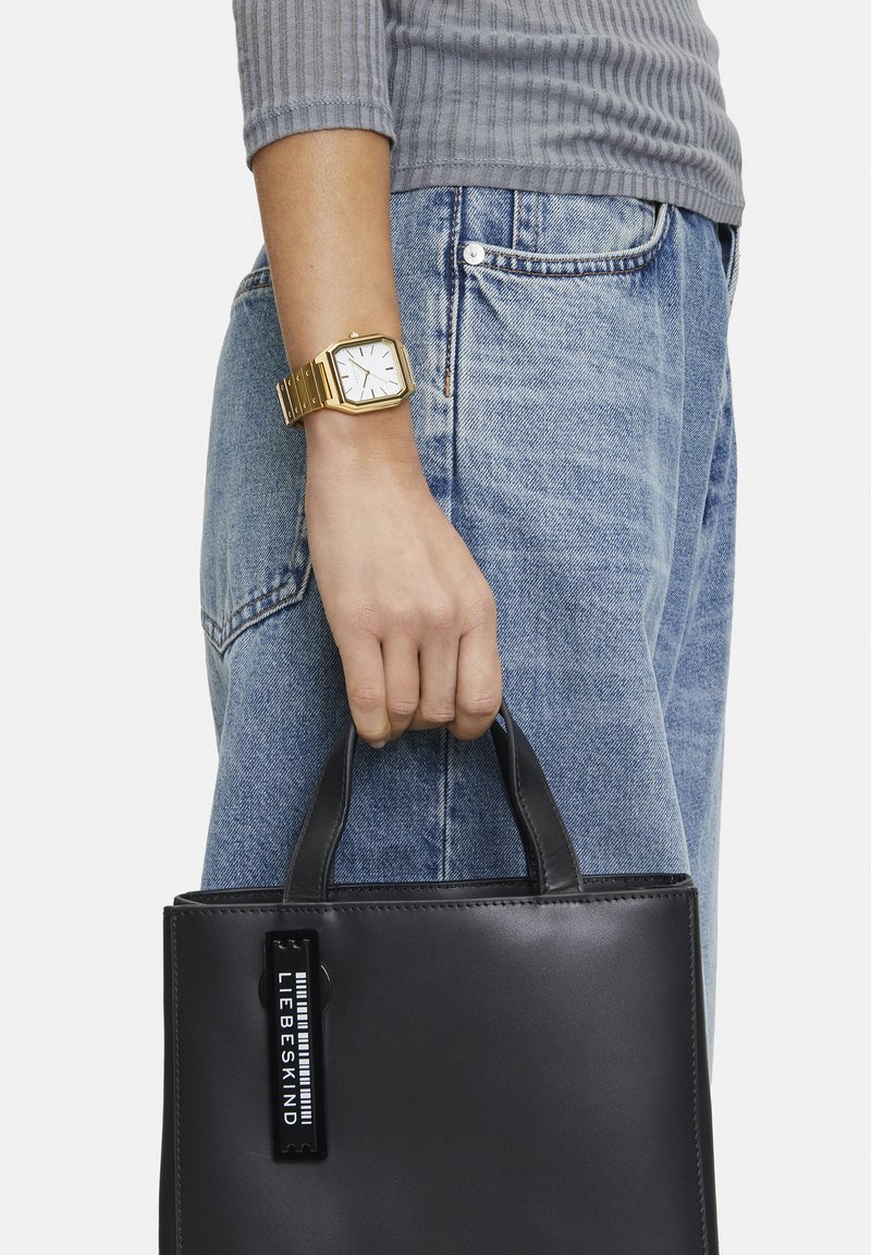 Svart läder tote bag med korta handtag och ett tag som står "LIEBESKIND." Kombination med ljusblå jeans och en grå ribbad topp. Guldur på handleden.