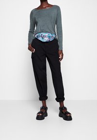 Harmaa pitkähihainen paita, mustat cargo-housut, joiden lahkeet on käärätty, ja monivärinen vyölaukku. Tukeva musta sandaalit soljilla.