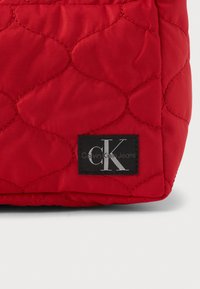 Κόκκινη επενδεδυμένη τσάντα από ύφασμα με μαύρη ετικέτα που φέρει την ένδειξη "Calvin Klein Jeans". Αξιοσημείωτα σχέδια ραφής και λεία υφή.