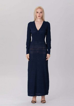 Femme debout portant une longue robe en maille bleu marine à manches longues et décolleté en V profond, associée à des talons ouverts noirs, sur un fond clair uni.