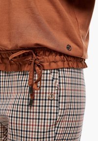 Brązowy sweter z marszczonym dołem i sznurkiem. Połączony z beżowymi spodniami w kratę, które mają czerwone i czarne akcenty oraz ozdobny guzik.