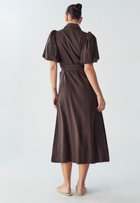 Robe brune midi avec des manches bouffantes, une taille ceinturée, un col et une texture lisse. La jupe tombe élégamment jusqu'aux chevilles.