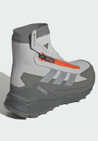Light gray high-top boots with Gore-Tex material, featuring an orange zipper, gray sole, and textured accents. Terrex branding visible.