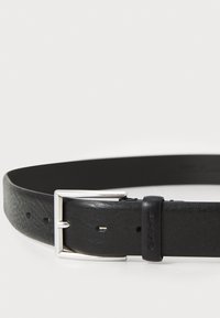 Cinturón de cuero negro con acabado texturizado, hebilla de metal plateado y un lazo de sujeción con marca en relieve. Ajustable con múltiples orificios.