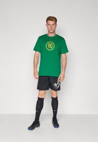 Grønt bomulds t-shirt med en gul cirkel og "90" grafik, sorte shorts, sorte knæhøje sokker og sorte fodboldstøvler med blå accenter.