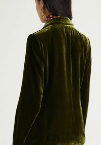 Veste en velours vert olive avec col cranté et coupe ajustée. Texture douce avec un léger éclat, mettant en valeur la brillance du tissu lisse.