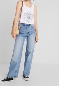 Débardeur blanc avec un graphique floral noir, associé à un jean large bleu clair et des baskets Converse noires. Sac en bandoulière marron.