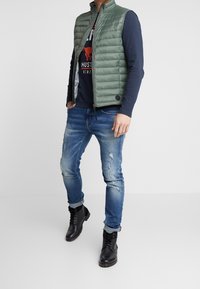 Grön vadderad väst med dragkedja, bärs över en marinblå långärmad skjorta med grafiskt tryck, parat med slitna blå jeans och svarta stövlar.