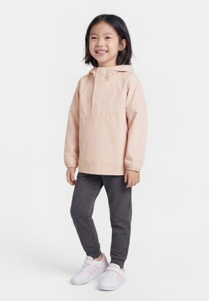 Χαμογελαστό παιδί που φοράει ανοιχτό ροζ μπουφάν με κουκούλα, σκούρο γκρι φόρμα και λευκά αθλητικά παπούτσια με ροζ ρίγες, στέκεται μπροστά σε λευκό φόντο.