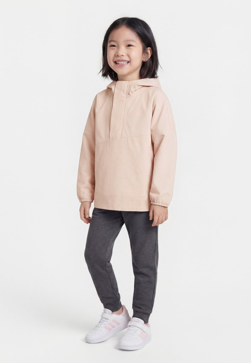 Χαμογελαστό παιδί που φοράει ανοιχτό ροζ μπουφάν με κουκούλα, σκούρο γκρι φόρμα και λευκά αθλητικά παπούτσια με ροζ ρίγες, στέκεται μπροστά σε λευκό φόντο.