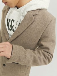 Hellbrauner Wollmantel mit Reverskragen und zwei Knöpfen, darunter ein graues Hoodie. Strukturiertes Material und taillierte Passform.