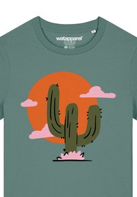 Groene t-shirt met een cactusafbeelding in het groen op een oranje cirkelvormige achtergrond, met roze wolken en een bloemachtige basis. Gemaakt van katoen.