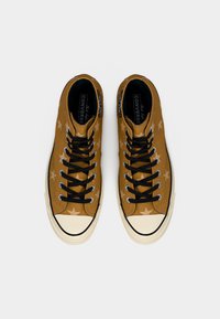 Pruunid kõrge säärega Converse tennised tähemustritega, mustade nauade ja valgete kummist sõrmeotsaga, vaadatuna ülevalt valgel taustal.