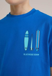 Kék pamut póló kerek nyakkal, három stilizált szörfdeszkával világoskék és fehér színekben, alatta a "BLUE RIDGE DENIM" felirattal.