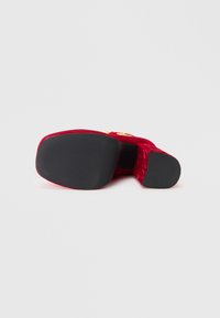 Chaussure à talons hauts en velours rouge avec une finition texturée, bout rond et semelle en caoutchouc noire. Présente des accents en métal doré sur la partie supérieure.