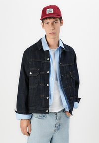 Denimjack met twee borstzakken, knoopsluiting en oranje stiksels. Ggedragen over een lichtblauwe gestreepte shirt en een witte t-shirt. Rode pet.