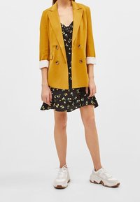 Senapsgul dubbelknäppt blazer över en svart blommig klänning. Klänningen har en volangkant, och vita sneakers fulländar outfiten.