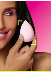 Rosa sminksvamp i form av en tårdropp som hålls av en hand, med en mjuk yta, mot en gradient rosa bakgrund. Klara accenter ramar in svampen.