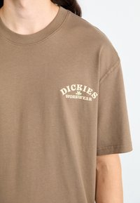 Brun bomull t-skjorte med rund hals, korte ermer og et fronttrykk som leser "Dickies Workwear" i krem og grønn.