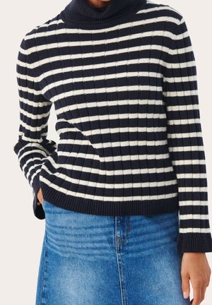 Person iført en marineblå og hvidstribet ribstrikket rullekravetrøje og en blå denim nederdel, med den ene hånd i lommen.