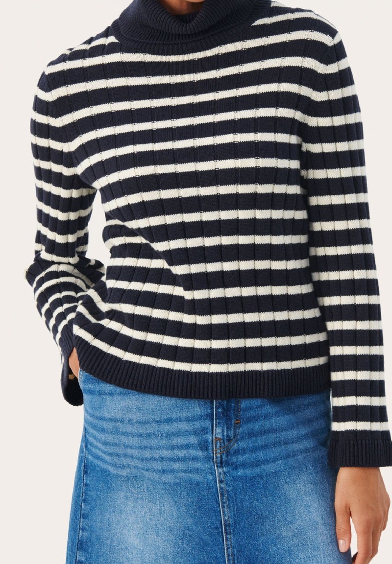 Personne portant un pull col roulé côtelé rayé bleu marine et blanc et une jupe en denim bleu, avec une main dans la poche.