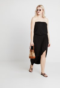 Svart axelbandslös klänning med en svängande silhuett och sidoklyfta, kombinerad med bruna sandaler och en liten vävd handväska.