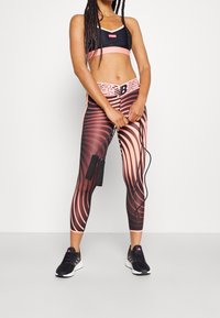 Reggiseno sportivo nero con dettagli rosa, leggings a fantasia di un rosa scuro con linee ondulate rosa chiaro e scarpe da ginnastica nere. In mano una corda per saltare.