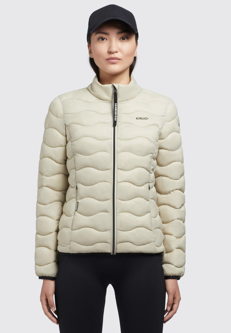 Jachetă puffer matlasată de culoare bej deschis, cu guler înalt, închidere cu fermoar, buzunare laterale și branding KHUJO. Textură netedă și un model subtil de valuri.