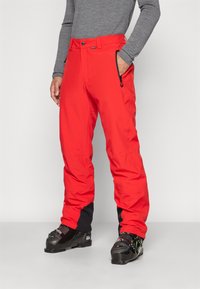 Pantalones de esquí rojos con un acabado texturizado, que presentan bolsillos con cremallera y acentos negros en los tobillos. Llevados con botas de esquí negras.
