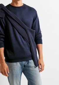 Marinblå sweatshirt i ett slätt material, med en ribbad krage och muddar, samt ett matchande stickat plagg som hänger över ena axeln.