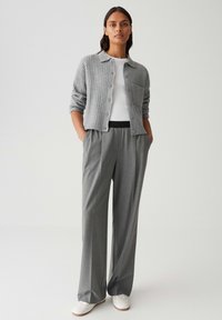 Donna in piedi con un cardigan grigio su una maglietta bianca, pantaloni larghi grigi, mani in tasca e sneakers bianche, su uno sfondo semplice.