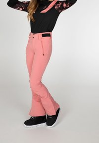Pantalon de ski rose en matière extensible, coupe ajustée, design évasé aux chevilles, avec poches zippées. Associé à des bottes de neige noires.