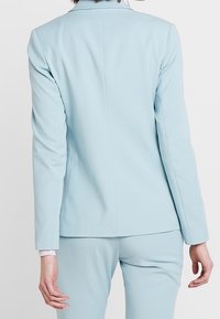 Blazer ajusté bleu clair en tissu lisse, avec un col classique et une coupe structurée, comprenant un détail de couture discret au dos.