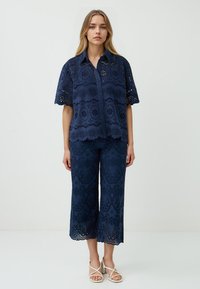 Una donna sta in piedi indossando una blusa ricamata blu navy e pantaloni a vita alta abbinati, insieme a sandali bianchi con cinturini, davanti a uno sfondo uniforme.