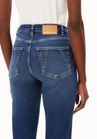 Jean bleu foncé à taille haute, coupe droite, avec des coutures contrastantes. Équipé d'un patch en cuir beige à l'arrière.