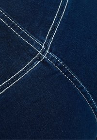 Denim materiál v tmavě modré barvě s bílým kontrastním prošitím. Obsahuje křížící se švy a hladkou texturu. Žádné viditelné vzory.