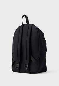 Mochila de tela negra con correas de hombro acolchadas y un asa de transporte en la parte superior, mostrada desde atrás sobre un fondo liso de color gris claro.