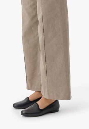 Zapatos negros de piel tipo mocasín con puntera redondeada y ribete elástico, combinados con pantalones de pierna ancha en un tono beis claro. Textura suave y diseño minimalista.