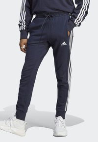 Temno modri Adidas trenirki s belimi črtami ob straneh, elastični pas, dvema stranskima žepoma in prilegajočimi se zapestji.