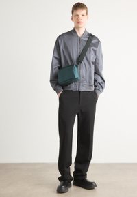 Giacca bomber grigia, pantaloni neri e scarpe nere voluminose. Borsa rettangolare teal con tracolla. Design elegante e contemporaneo.