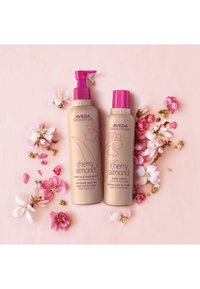 Twee Aveda cherry almond verzorgingsproducten in beige flacons met roze pompen, omringd door roze kersenbloesembladeren op een lichtroze achtergrond.