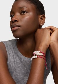 Tres pulseras con perlas blancas y cuerdas trenzadas rojas, llevadas en una muñeca, mostrando texturas suaves y acentos de pequeños cristales.