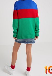 Person som bär en färgblockströja i blått, rött och grönt, en fransad denimkjol, röda strumpor och vita sneakers, stående med ryggen mot kameran.